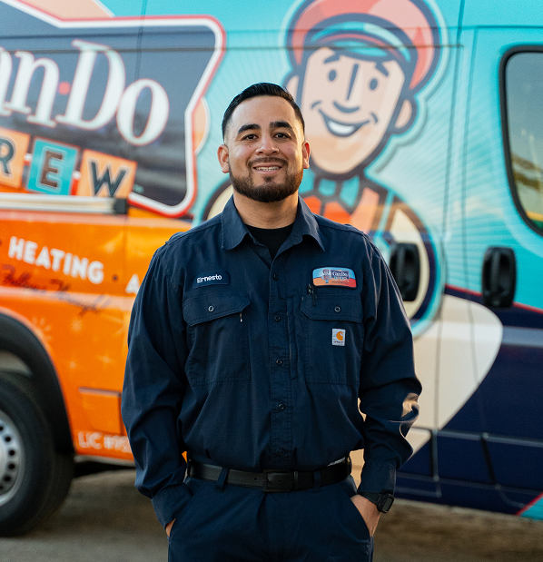 Male technician in a dark blue uniform stands with hands in pockets in front of a colorful service van, smiling at the camera (name badge reads Ernesto).