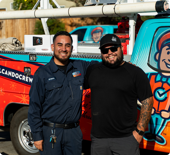 Two smiling men posing in front of a colorful tow truck with a cartoon mascot; one in a navy work uniform, the other in a black t-shirt and cap.
