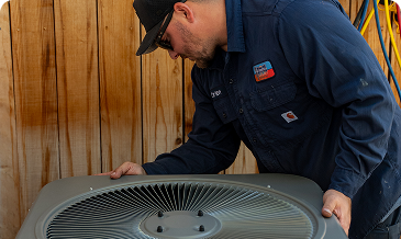 HVAC technician in a blue shirt and cap inspecting an outdoor air conditioner condenser unit.