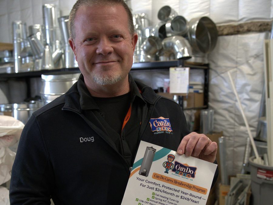Doug, a plumbing technician, stands in a metal workshop holding a Can Do Crew membership flyer.