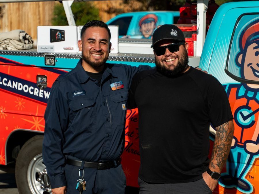 Two men smiling and posing in front of a bright, cartoon-themed service truck; one in a navy uniform with a name tag.