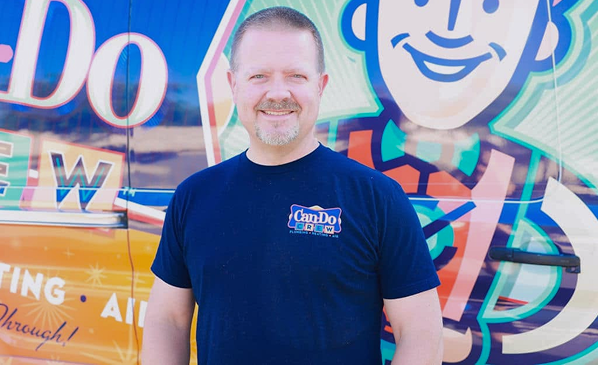 Smiling man wearing a blue Can Do Crew T-shirt stands in front of a brightly painted van with a large cartoon mascot image behind him.
