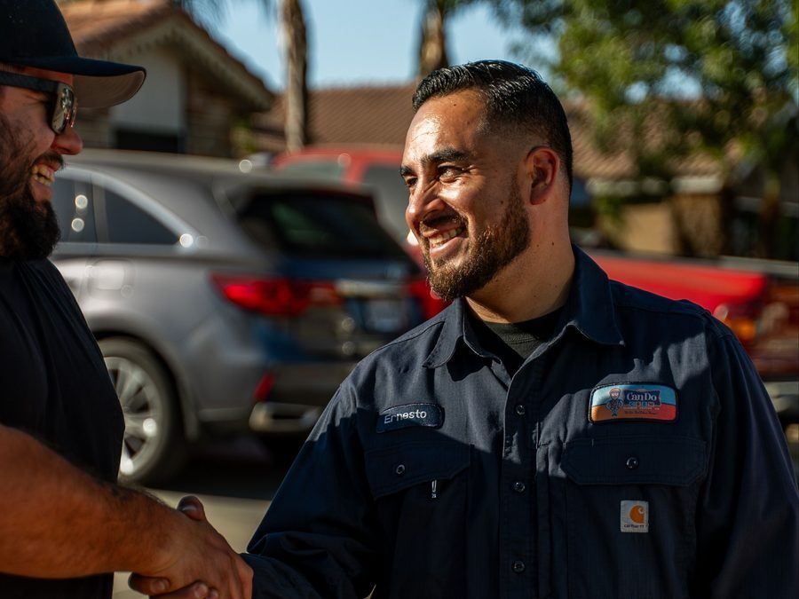 Two men shake hands outdoors; one man in a dark work shirt named Ernesto smiles at the other.