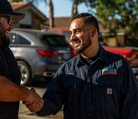 Two men shake hands outdoors, one in a dark work shirt with a name badge, smiling at the moment of greeting.