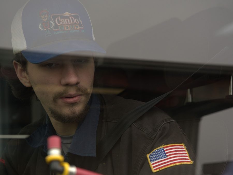 Young man in a CanDo Crew cap peers through a window, with an American flag patch on his brown jacket sleeve
