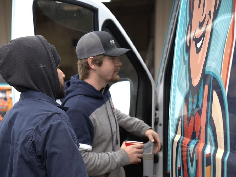 Two men stand by a colorful mural on the side of a van, one holding a red cup and looking at the artwork.