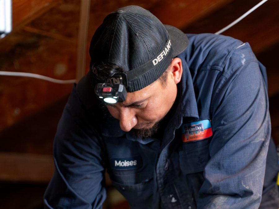 Tradesman moises wearing a headlamp and Defiant cap, concentrating on a task in a dusty workshop attic.
