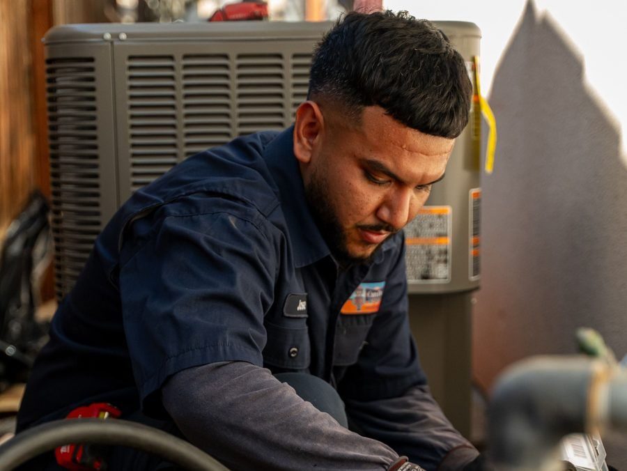HVAC technician in a navy shirt crouches by an outdoor air conditioner condenser, inspecting the unit.