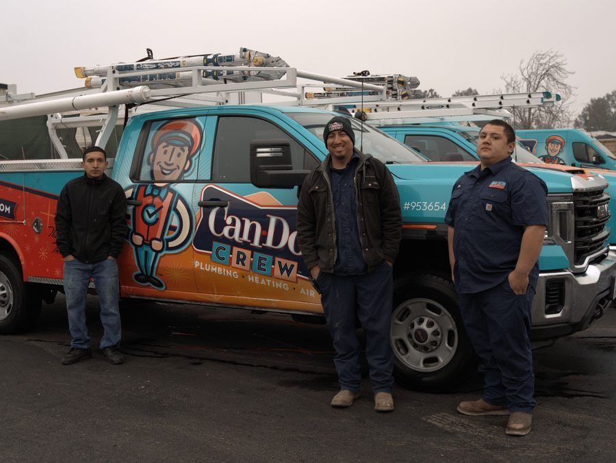 Three technicians stand in front of a teal and orange Can-Do Crew plumbing van with ladders on the roof on a cloudy day.