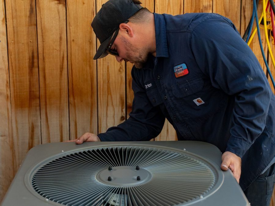 HVAC technician in a blue uniform inspecting an outdoor air conditioner unit near a wooden fence.