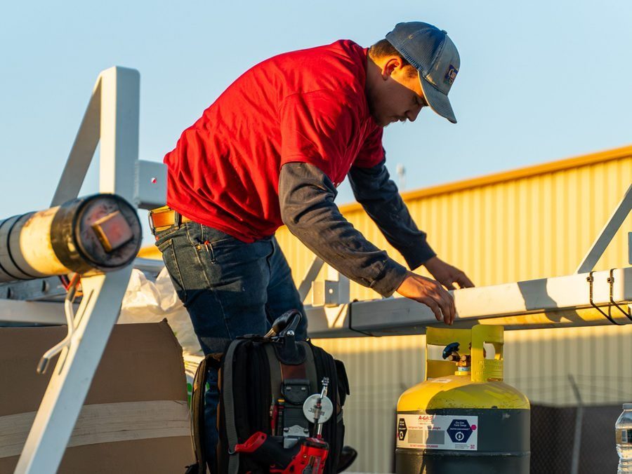 Maintenance worker in a red shirt and cap adjusting a metal beam on a rooftop beside a yellow propane tank and tools in a bag below him.