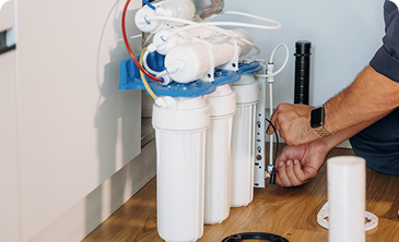 Technician installing or maintaining a multi-filter water purifier under a kitchen sink, with white filter housings and blue mounting.