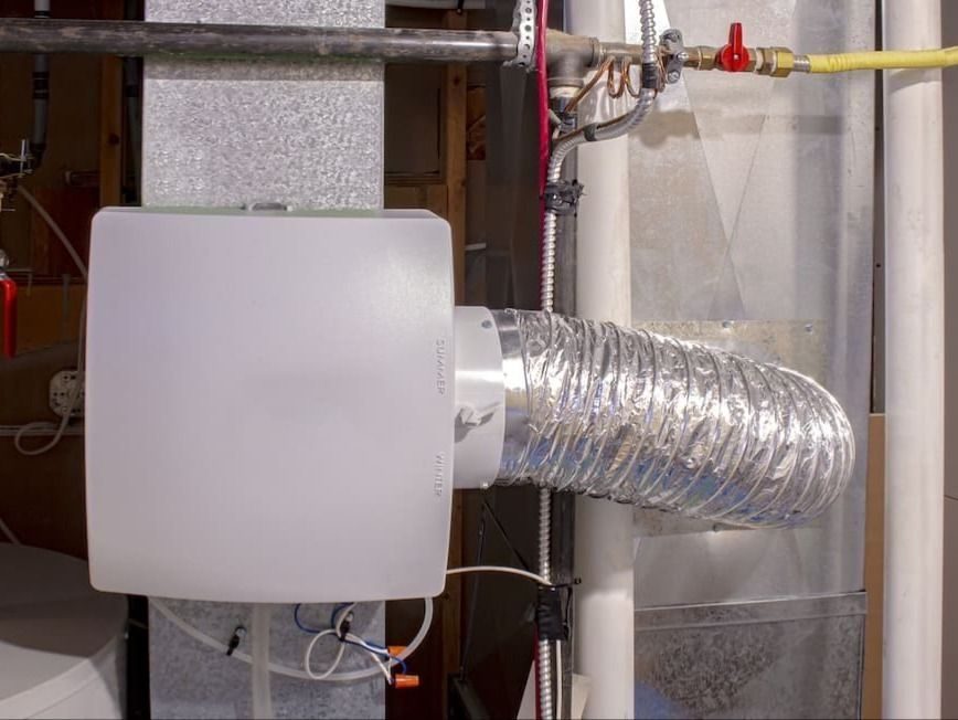 White rectangular HVAC unit connected to a flexible aluminum foil duct, part of an exhaust/vent system in a basement with wires nearby.
