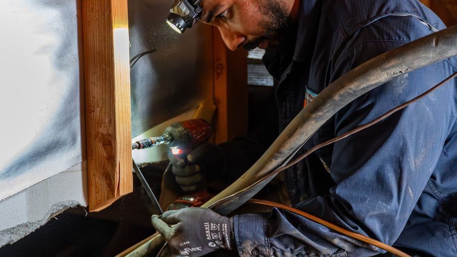 HVAC technician installing an AC system in an attic, working on refrigerant lines with a power drill.